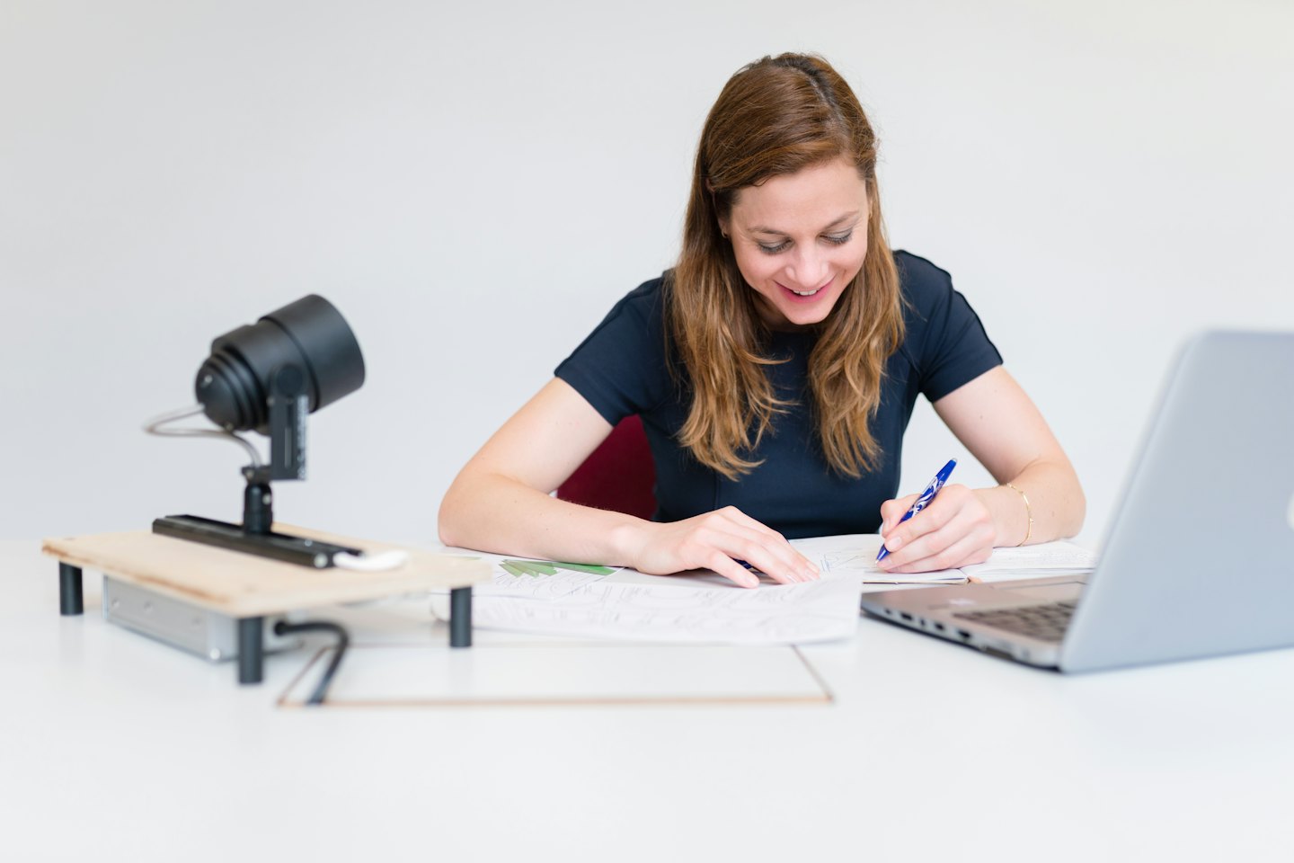 Žena u stolu se usmívá a píše perem, poblíž je notebook.